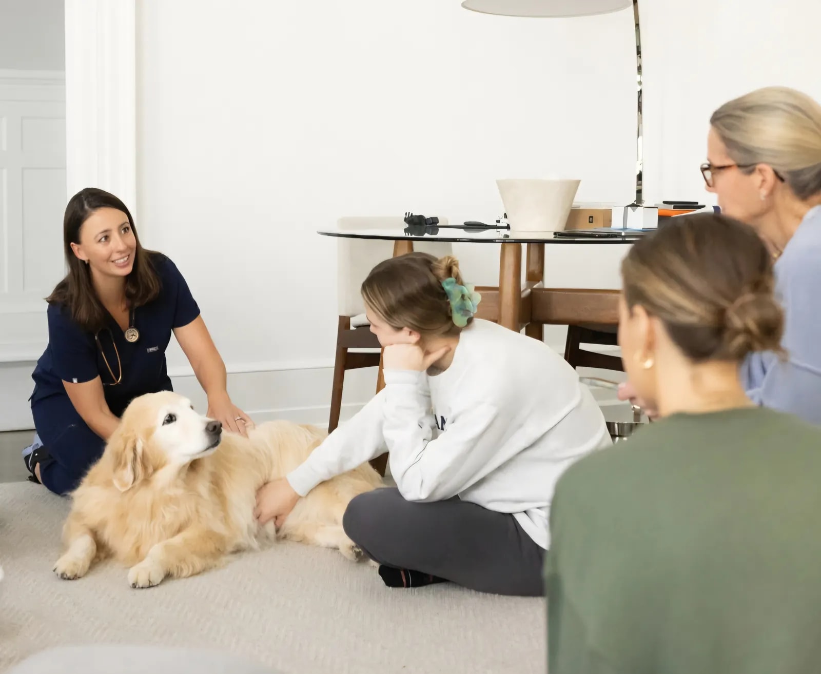 Dr. McCaffrey kneels beside a golden retriever lying on the floor while a family sits around watching and petting the dog