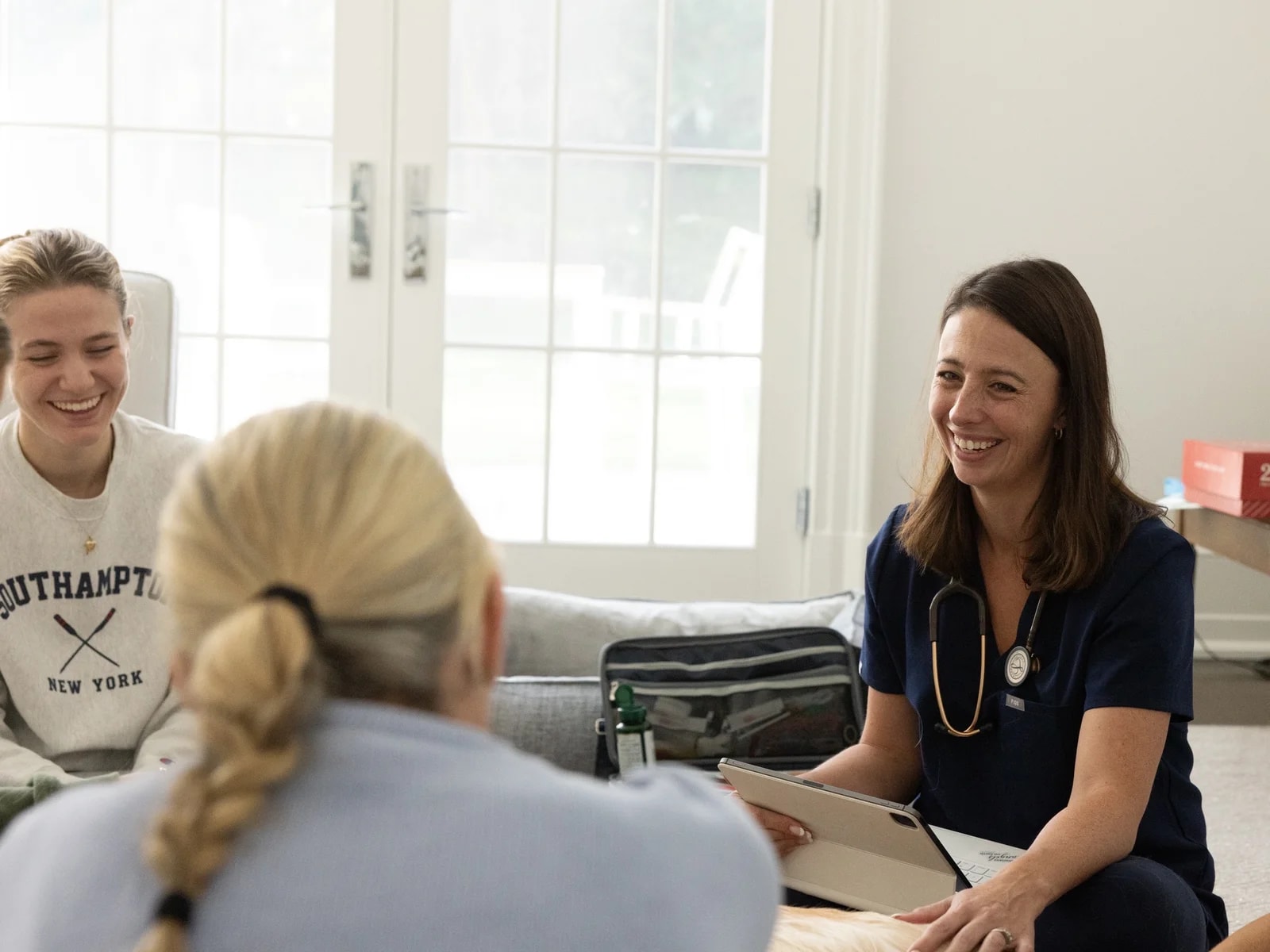 A smiling veterinary nurse holding a tablet while talking with a family and their dog in a bright living room